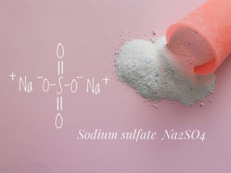 A pink scoop spills fine white powder—sodium sulfate (Na₂SO₄)—onto a soft pink surface, with its chemical structure elegantly illustrated beside it. The scene evokes cleanliness and precision, representing the high standards of Park Avenue Laundry, Bemidji, Minnesota 56601, where science meets spotless service.